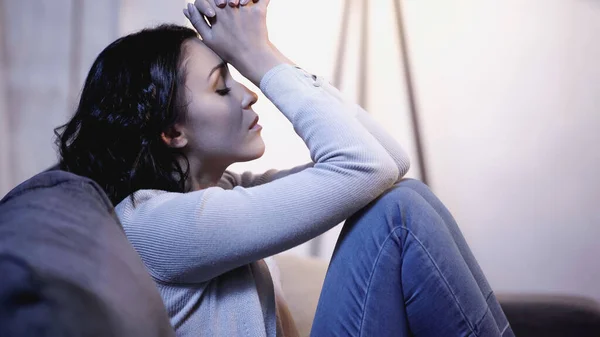 Femme déprimée en vêtements décontractés assis sur le canapé avec les yeux fermés et tenant les mains serrées près de la tête à la maison — Photo de stock