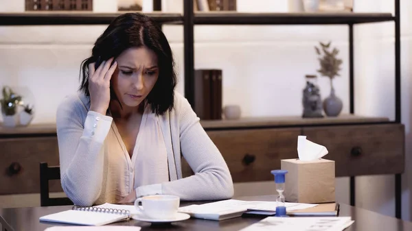 Mujer estresada sentada en la mesa con papeles y taza de café y sosteniendo la mano cerca de la cabeza en casa - foto de stock