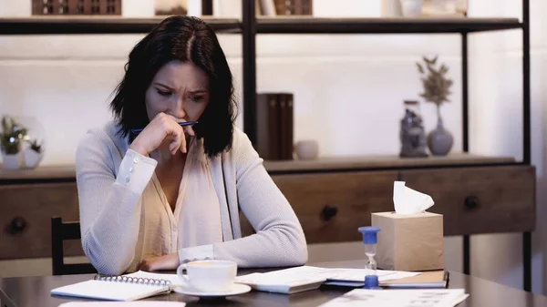 Worried woman sitting on table with cup of coffee, calculator, notebooks and holding pen in hand near face at home — Stock Photo