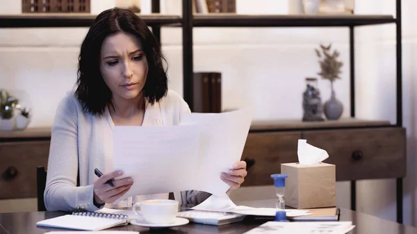 Femme inquiète assise sur la table et lisant des documents avec stylo à la main à la maison — Photo de stock