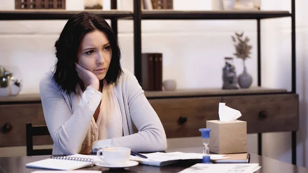 Depressed woman sitting on table with papers and cup of coffee and holding hand near face at home — Stock Photo