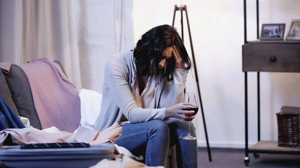 Подавленная женщина сидит на диване с бокалом красного вина рядом с мужской одеждой и держит руку рядом с головой дома — стоковое фото
