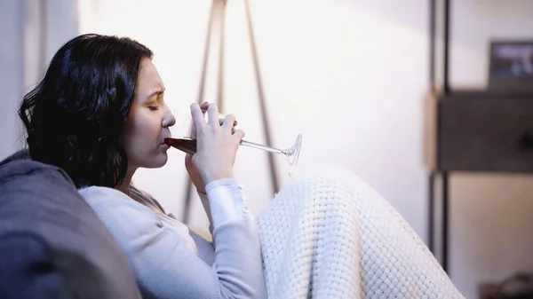 Femme bouleversée couverte de couverture assise sur le canapé et buvant du vin rouge du verre à la maison — Photo de stock