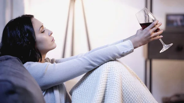 Donna sconvolta coperta di coperta seduta sul divano con bicchiere di vino in mani tese a casa — Foto stock