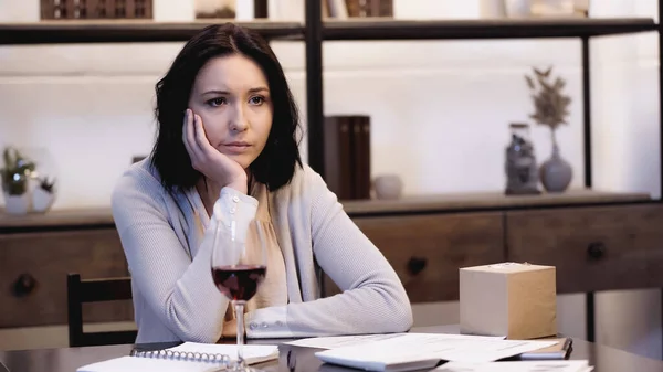 Depressed woman sitting on table with papers and glass of red wine and holding hand near face at home — Stock Photo