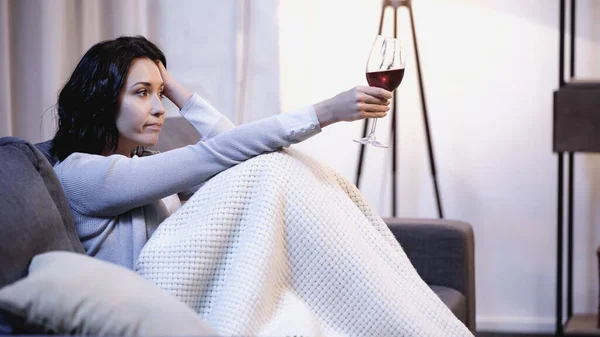 Femme bouleversée couverte de couverture assise sur le canapé avec un verre de vin et tenant la main près de la tête à la maison — Photo de stock