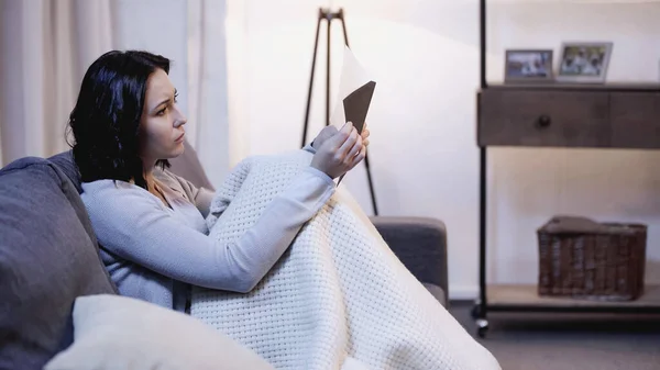 Upset woman covered in blanket sitting on sofa and looking at envelope in hand at home — Stock Photo