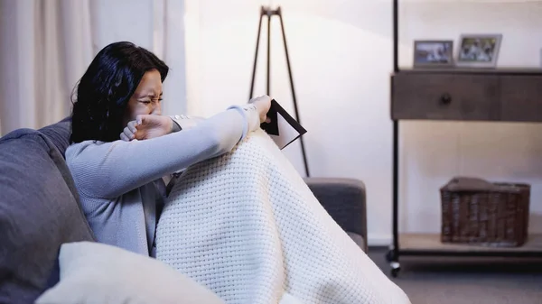 Mujer llorando sentada en el sofá, sosteniendo el sobre y cubriendo la cara con la mano en casa - foto de stock