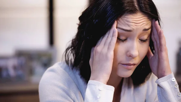 Upset woman sitting with closed eyes and holding hands near head at home — Stock Photo