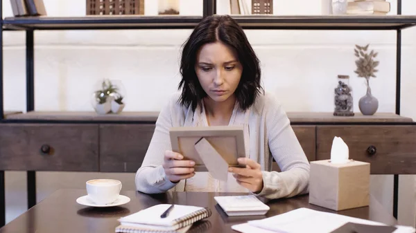 Upset woman sitting on table and holding photo frame in hands at home — Stock Photo