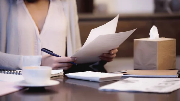 Partial view of woman sitting on table with cup of coffee and holding documents in hands at home — Stock Photo