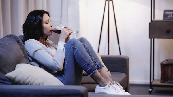 Femme déprimée solitaire en vêtements décontractés assis sur le canapé et boire du vin rouge du verre à la maison — Photo de stock
