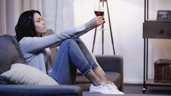 Депрессивная женщина в бежевом свитере и джинсах сидит на диване и держит стакан красного вина в протянутых руках дома — стоковое фото