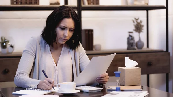 Donna preoccupata seduta sul tavolo con tazza di caffè op, in possesso di documenti e la scrittura in notebook a casa — Foto stock