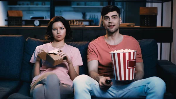 Happy man holding bucket with popcorn and clicking channel near girlfriend holding cardboard box with chinese food — Stock Photo