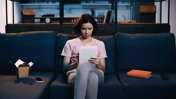 Молода жінка використовує цифровий планшет, сидячи на дивані біля смартфона, книжкової та картонної коробки з китайською їжею — стокове фото