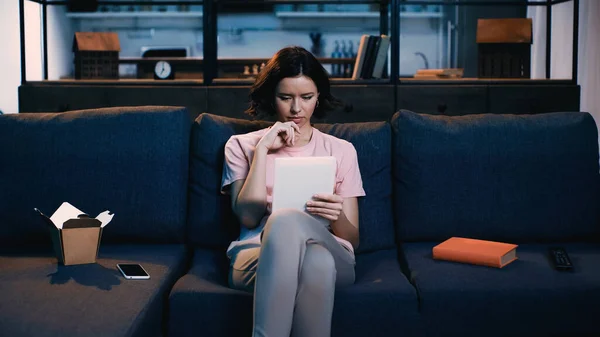 Brünette Frau mit digitalem Tablet, während sie auf dem Sofa neben Smartphone, Buch und Karton mit Lebensmitteln sitzt — Stockfoto