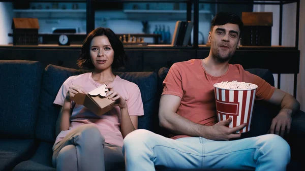 Cheerful man holding bucket with popcorn and watching comedy movie with girlfriend holding cardboard box with chinese food — Stock Photo