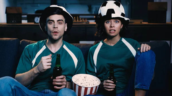 Fans de football excités dans des chapeaux de ventilateur tenant des bouteilles de bière et regardant le championnat dans le salon — Photo de stock