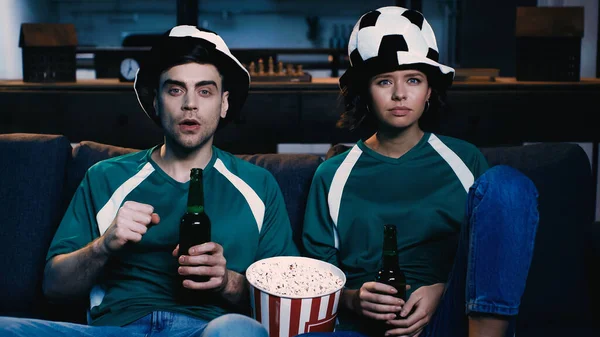 Fãs de futebol em chapéus de fãs segurando garrafas de cerveja e assistindo campeonato no sofá cerrado punho — Fotografia de Stock