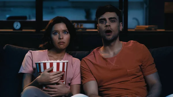 Woman holding popcorn bucket and watching scary movie with shocked boyfriend in evening — Stock Photo