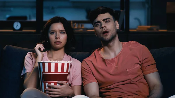 Displeased woman holding popcorn bucket and watching movie with surprised boyfriend — Stock Photo