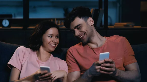 Alegre hombre y mujer sosteniendo los teléfonos celulares y sonriendo en la sala de estar moderna - foto de stock