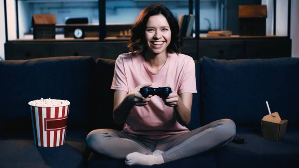 KYIV, UCRAINA - GIUGNO 09, 2021: donna gioiosa che tiene il joystick e gioca al videogioco vicino al secchio di popcorn sul divano — Foto stock