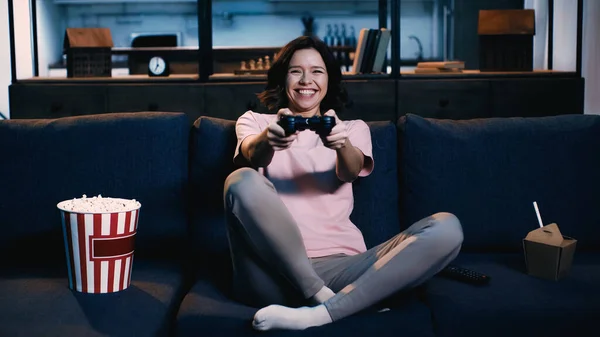 KYIV, UKRAINE - JUNE 09, 2021: happy woman holding joystick and playing video game near popcorn bucket on sofa — Stock Photo