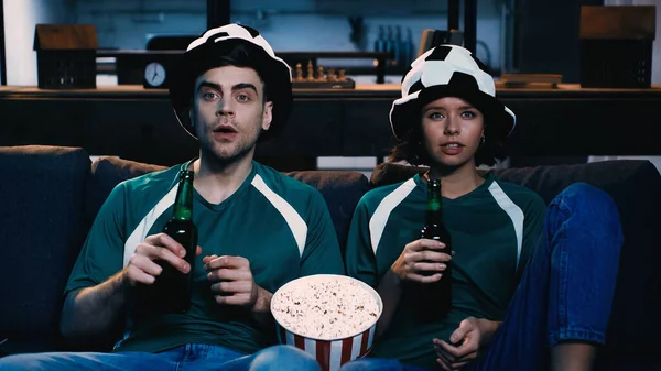 Напряженные футбольные болельщики в шляпах болельщиков, держащие бутылки пива и смотрящие чемпионат возле ведра попкорна — стоковое фото
