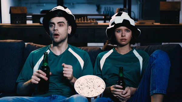Sorprendido aficionado al fútbol en abanico sombrero y mujer en camiseta verde sosteniendo botellas de cerveza y viendo el campeonato - foto de stock