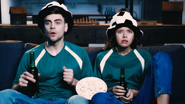 Tensed man and woman in fan hats holding bottles of beer and watching championship in living room — Stock Photo