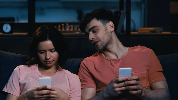 Curioso hombre mirando a la mujer mensajes de texto en el teléfono inteligente en la sala de estar moderna - foto de stock