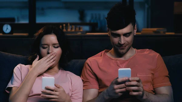 Zufriedene Männer und müde Frauen beim SMS-Schreiben auf Mobiltelefonen im modernen Wohnzimmer — Stockfoto