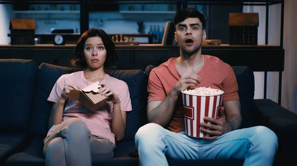 Schockierter Mann schaut Film und hält Popcorn-Eimer neben Frau, die mit chinesischem Essen in Karton sitzt — Stockfoto