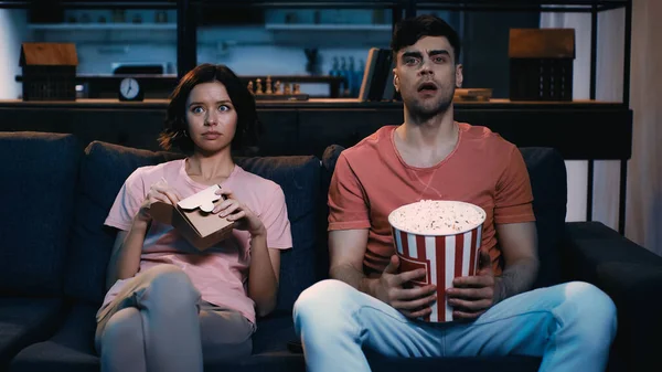 Überraschter Mann schaut Film und hält Popcorn-Eimer neben Frau, die mit Pappschachtel sitzt — Stockfoto