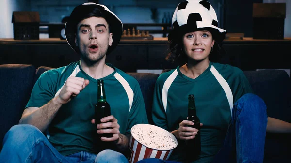 Worried couple in fan hats watching championship and holding bottles of beer near popcorn bucket in living room clenched fist — Stock Photo