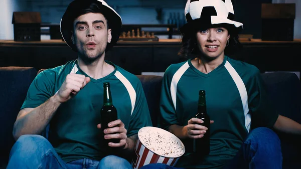 Casal estressado em chapéus de ventilador segurando garrafas de cerveja e assistindo campeonato perto de balde com pipocas — Fotografia de Stock