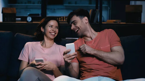 Hombre riendo con la mujer y la celebración de teléfonos inteligentes en la sala de estar moderna - foto de stock