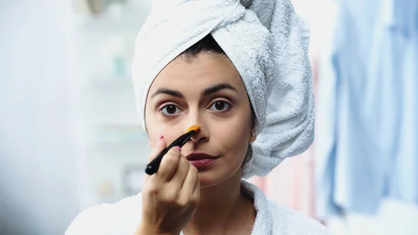 Jovem com cabeça embrulhada em toalha aplicando fundação no nariz com escova cosmética no quarto — Fotografia de Stock