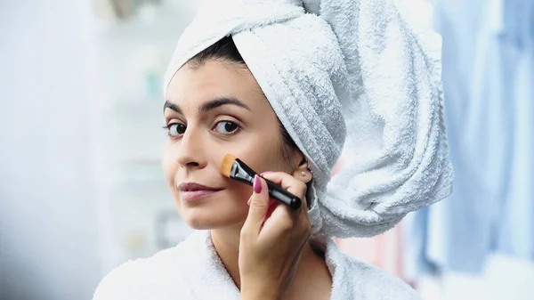 Mujer joven con la cabeza envuelta en toalla aplicando la base en la mejilla con cepillo cosmético en el dormitorio - foto de stock