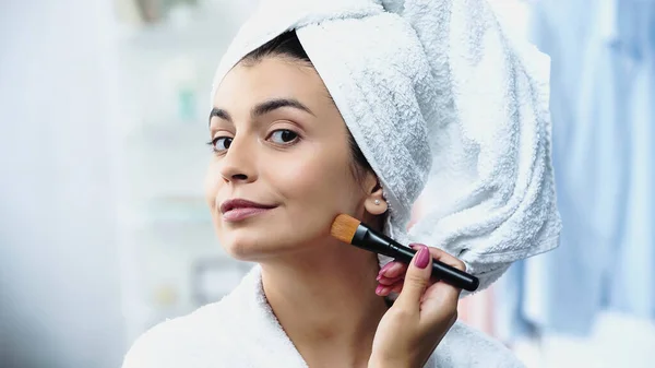 Mujer joven con la cabeza envuelta en toalla aplicando la base en la cara con cepillo cosmético en el dormitorio - foto de stock