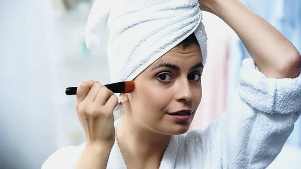 Jovem bonita com cabeça embrulhada em toalha aplicando fundação no rosto com escova cosmética no quarto — Fotografia de Stock