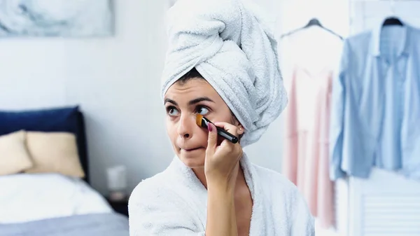 Young woman with head wrapped in towel applying face foundation under eyes with cosmetic brush in bedroom — Stock Photo