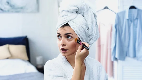 Jovem mulher em roupão de banho com cabeça embrulhada em toalha aplicando fundação no rosto com escova cosmética no quarto — Fotografia de Stock