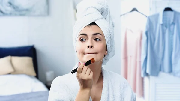 Jovem mulher em roupão de banho com cabeça embrulhada em toalha grimacing e aplicando fundação rosto no quarto — Fotografia de Stock