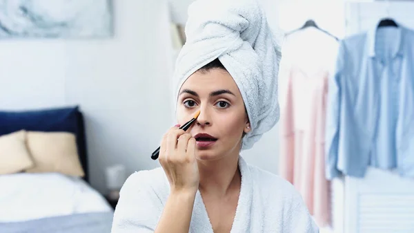 Giovane donna con bocca aperta e testa avvolta in asciugamano applicando fondazione viso in camera da letto — Foto stock