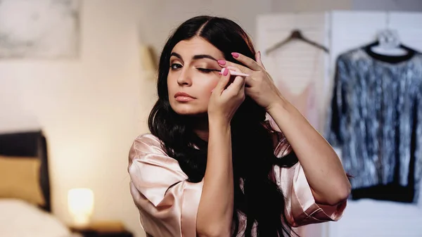 Concentrada jovem mulher em seda rosa peignoir aplicando eyeliner no quarto — Fotografia de Stock