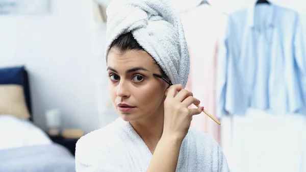 Focused young woman with head wrapped in towel styling eyebrow with cosmetic brush in bedroom — Stock Photo