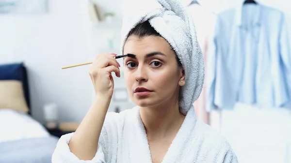 Jovem mulher em roupão de banho com cabeça envolta em sobrancelha estilo toalha com escova no quarto — Fotografia de Stock